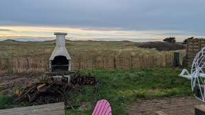 Un entorno rústico al aire libre con una chimenea de piedra, una valla de madera y un paisaje cubierto de césped bajo un cielo nublado.