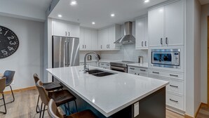 A modern kitchen with white cabinetry, stainless steel appliances, a large island with bar stools, and a wall clock.