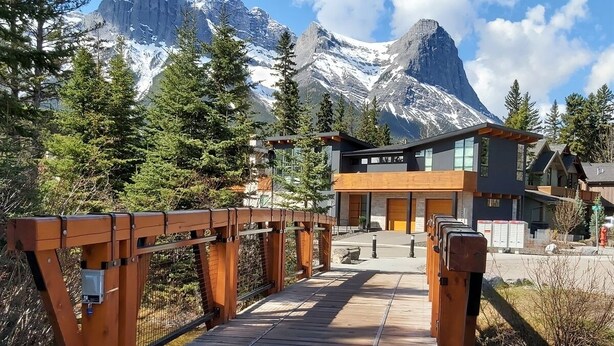 A wooden bridge leading to a modern building with large windows, surrounded by snow-capped mountains and evergreen trees.