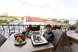 Un balcón con una mesa dispuesta para el desayuno, que incluye un plato de frutas y un sándwich, y vistas a un pueblo con edificios y un monumento a lo lejos.
