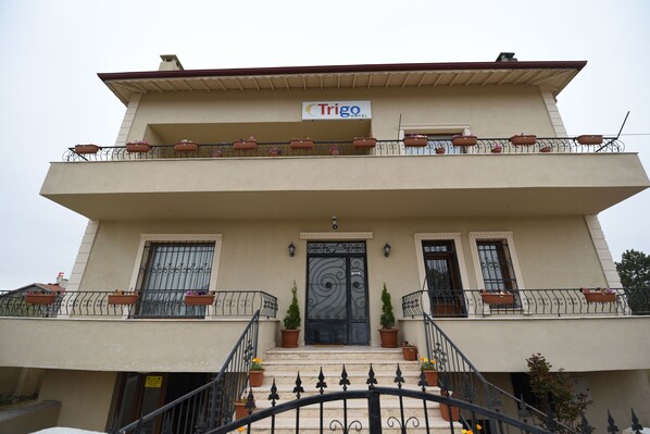 Un edificio de dos plantas con un balcón en el piso superior, un letrero que dice "Trigo Hotel" sobre la entrada, y macetas con plantas en las barandillas del balcón.