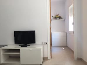 A room with a TV on a white desk, a white wardrobe, and a shelf with decorative items.