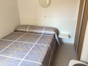A small, neatly made bed with a checkered bedspread and pillows, a white bedside table, and a wall-mounted light fixture.