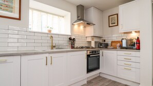 A modern kitchen with white cabinets, a stainless steel range hood, a built-in oven, and a window with blinds. - Bay Cottage No 5 (Morecambe)