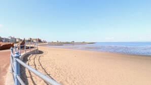 A sandy beach with a metal railing, buildings in the background, and a clear sky. - Bay Cottage No 5 (Morecambe)