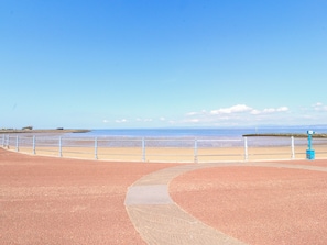 A paved walkway with a metal railing, leading to a beach with a clear view of the sea and distant land. - Bay Cottage No 5 (Morecambe)