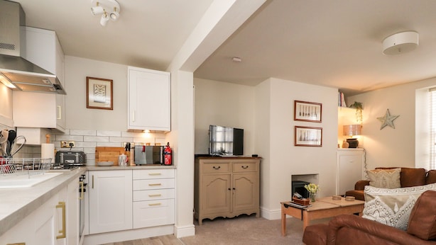 A modern kitchen with white cabinets, a dining area with a brown leather sofa, and a fireplace. - Bay Cottage No 5 (Morecambe)