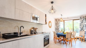 Una cocina moderna con microondas integrado, horno y lavaplatos. Hay una zona de comedor con una mesa y sillas, y vistas al exterior a través de amplios ventanales.