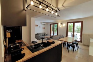 A modern kitchen with a central island, a dining area with a table and chairs, a sofa, and a large window with a view of greenery.
