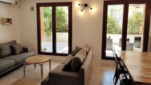 Ein Wohnzimmer mit einer Couch, einem kleinen runden Tisch und einem Essbereich mit einem Holztisch und Stühlen. Große Schiebeglasfenster führen zu einer Außenpatio mit Möbeln.