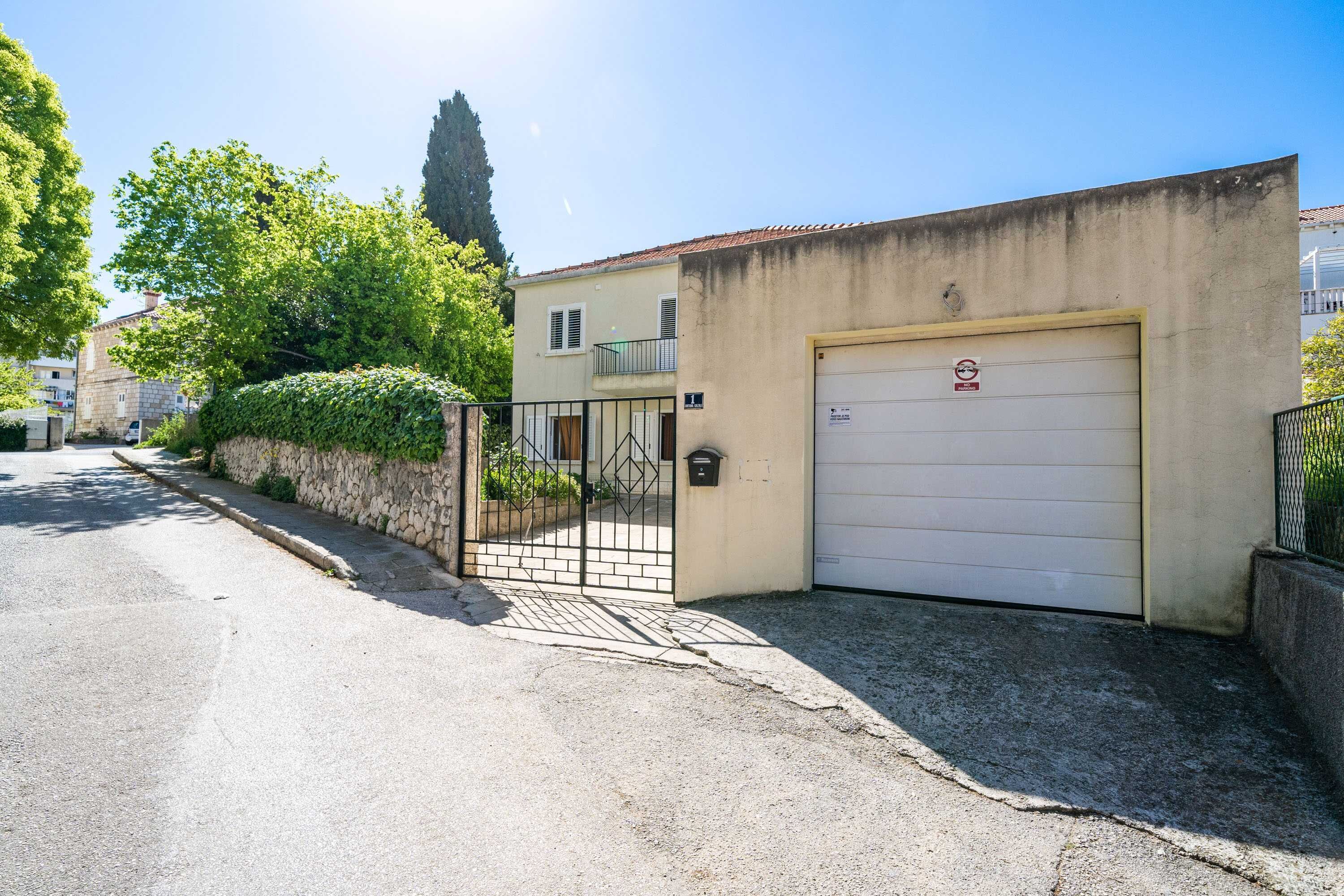A residential street with a house featuring a garage door, a stone wall, and a metal gate.