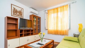 A living room with a green sofa, wooden entertainment unit, TV, and a window with orange curtains. - Apartment Kos (Dubrovnik)