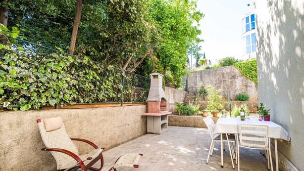 An outdoor patio with a table and chairs, a barbecue grill, and a seating area with a cushioned chair and a bench. - Apartment Kos (Dubrovnik)