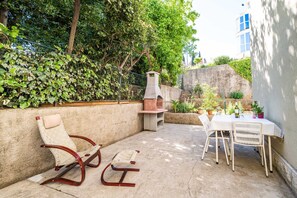 An outdoor patio with a table and chairs, a barbecue grill, and a seating area with a cushioned chair and a bench. - Apartment Kos (Dubrovnik)