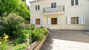 A two-story building with a balcony on the second floor, a concrete pathway leading to the entrance, and a garden area with various plants and flowers. - Apartment Kos (Dubrovnik)