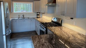 A modern kitchen with grey cabinets, a stainless steel refrigerator, a gas stove, and a window providing a view of trees. - Beautiful Mink Lake, Close to Algonquin Park (Lake St. Peter)