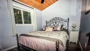 A bedroom with a wooden ceiling, a bed with a metal headboard, a nightstand, a window with curtains, and a view of trees outside. - Beautiful Mink Lake, Close to Algonquin Park (Lake St. Peter)