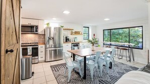 Eine moderne Küche mit Edelstahlgeräten, ein Essbereich mit Holztisch und -stühlen sowie große Fenster mit Blick auf Grünflächen.