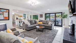 Ein modernes Wohnzimmer mit einem grauen Ecksofa, einem Couchtisch und einem Kamin. Im Hintergrund befindet sich eine Küche mit Essbereich.
