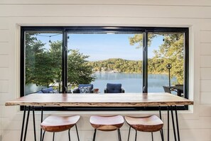 A dining area with a large window overlooking a lake, a wooden table, and three bar stools.