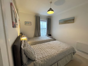 A bedroom with two beds, a window with curtains, a hanging light fixture, and a wall-mounted painting. - Coastal Cottage at Sea Palling dog friendly (Sea Palling)