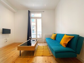 En stue med en tealgrøn sofa, et træsofabord, en gul tæppe og et tv ophængt på væggen.