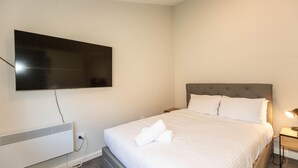 Una camera d'albergo con un letto, una TV a schermo piatto montata a parete, un comodino con una lampada e un termosifone.