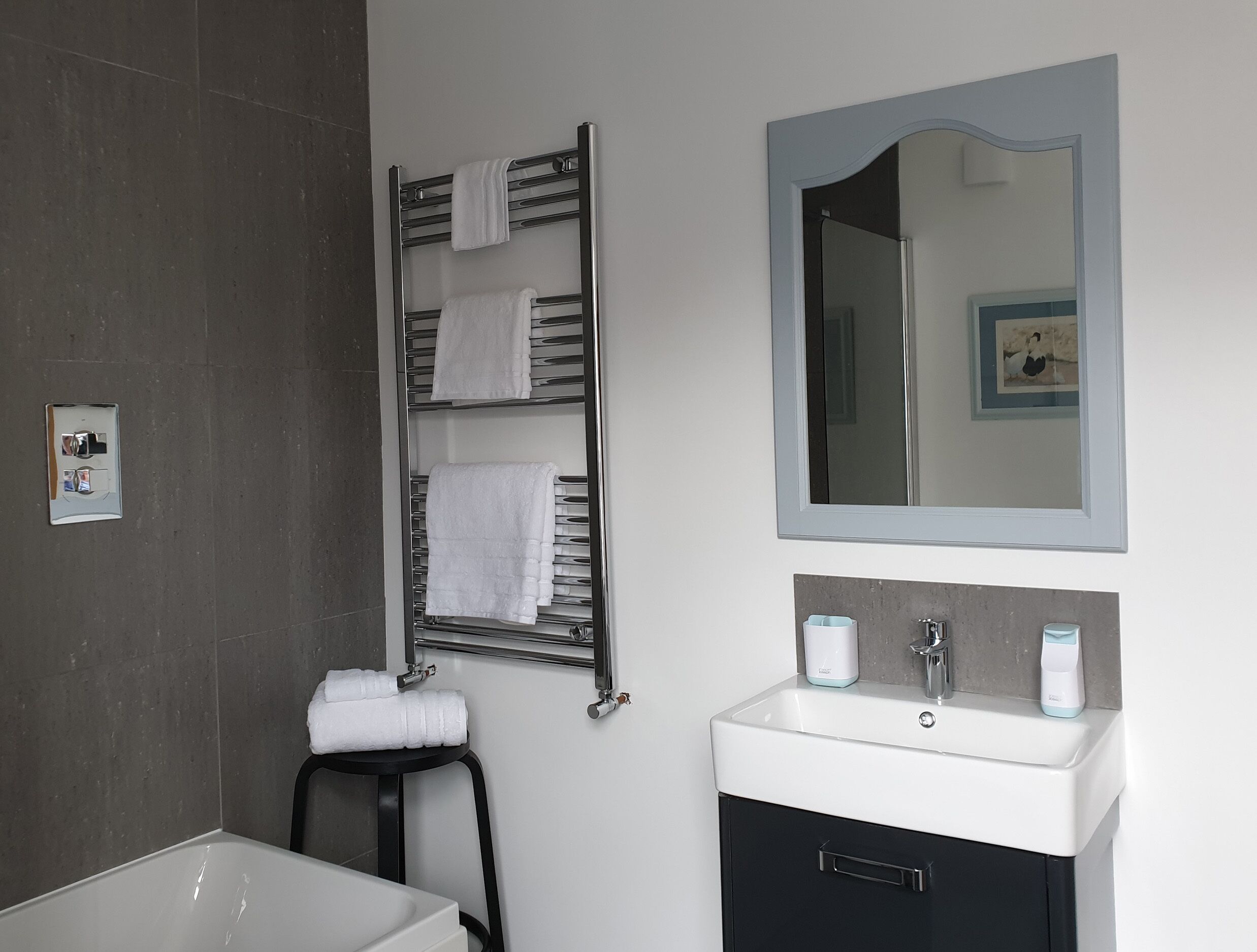 A modern bathroom with a bathtub, a towel rack with multiple towels, a sink with a mirror above it, and a toilet paper holder.