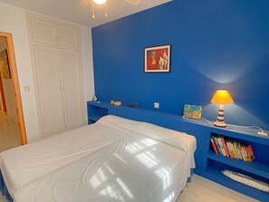 A bedroom with a bed, a blue wall, a painting, a lamp, and a bookshelf. - Ample duplex frontline to the Med (La Manga del Mar Menor)