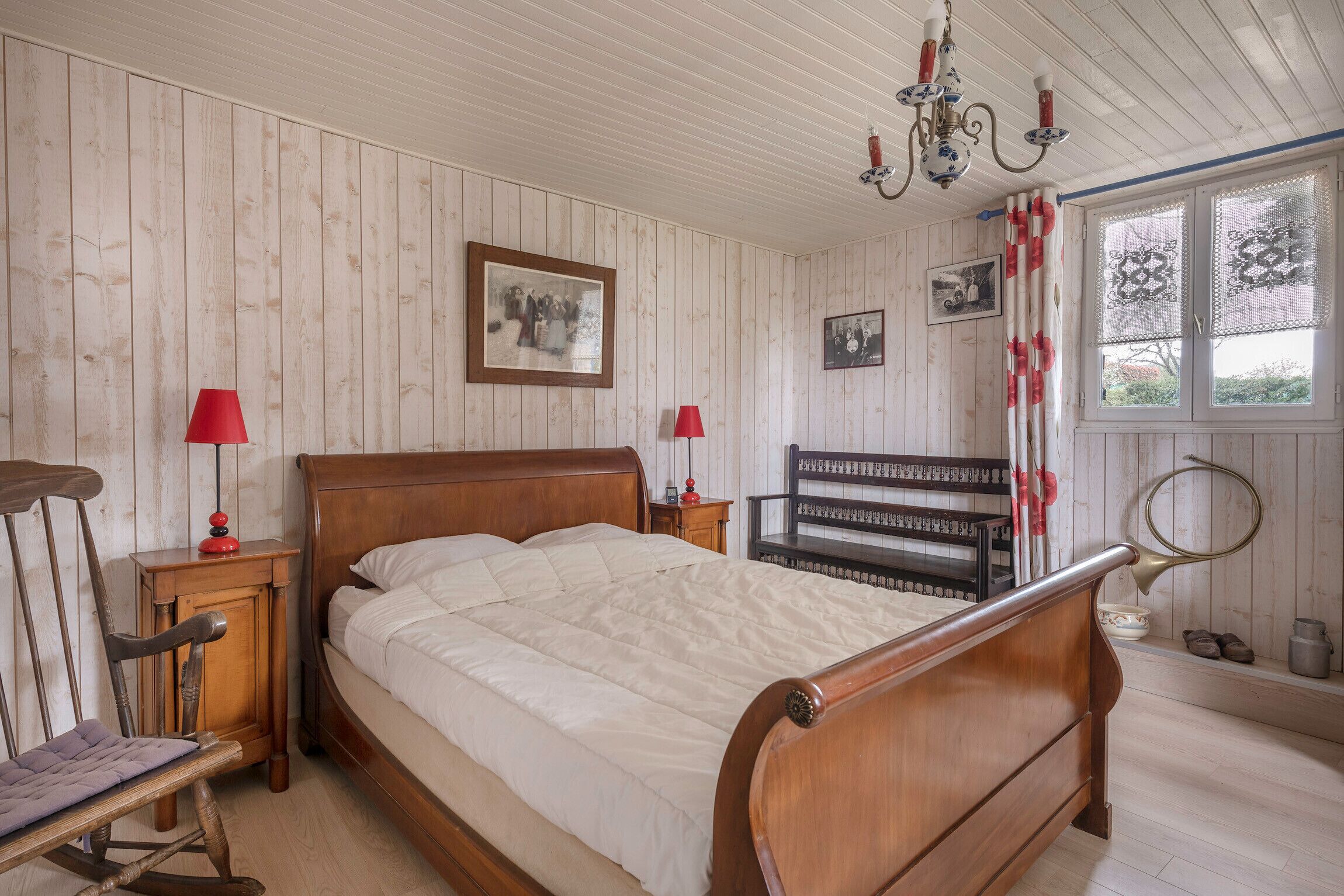 Un dormitorio con una cama de madera, dos mesitas de noche, una lámpara roja, un candelabro y una ventana con cortinas de encaje.