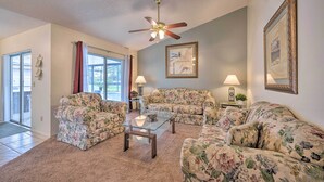 Sala de estar con sofás de estampado floral, una mesa de centro de vidrio y un ventilador de techo. Hay dos lámparas de pared y un cuadro en la pared.