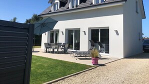 A modern two-story house with a sloped roof, large sliding glass doors, and a patio area with outdoor furniture. - Villa Rubis, an ecological house with modern comfort (Varengeville-sur-Mer)