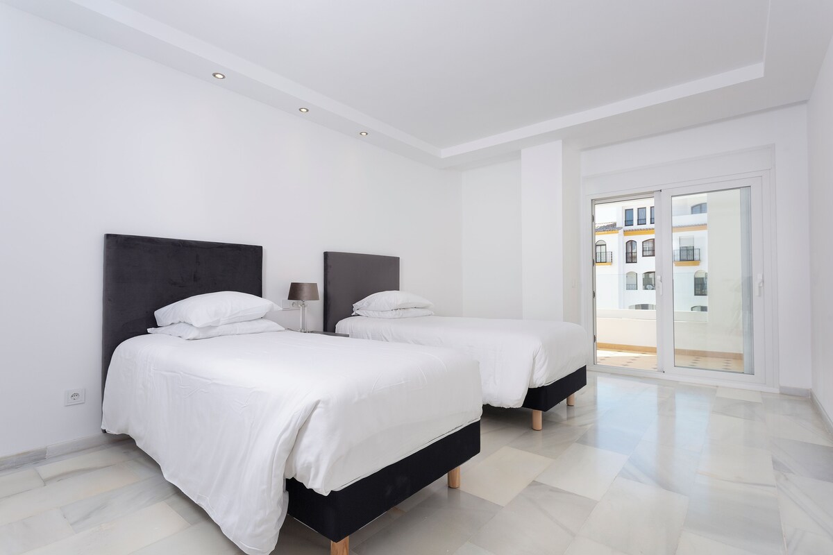 A modern bedroom with two beds, a large window, and a view of buildings outside.