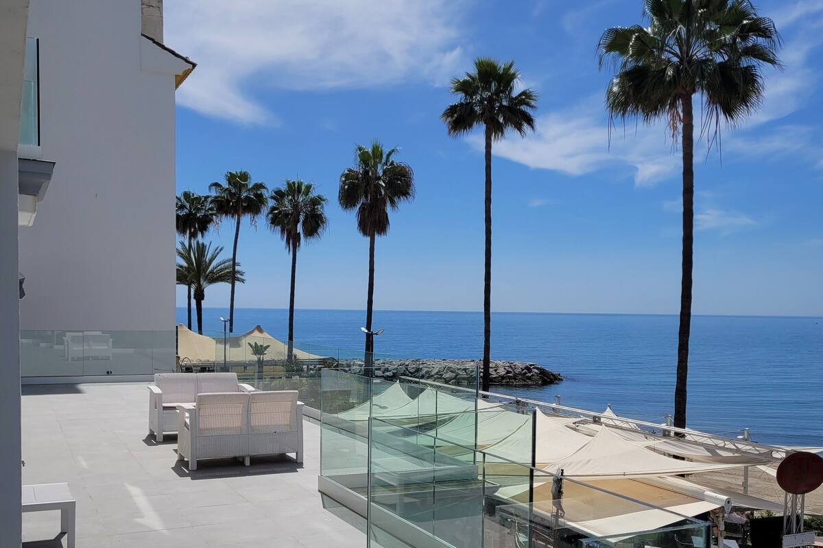 Una terraza en la azotea con barandas de vidrio, muebles de mimbre blanco y palmeras con vista al mar.