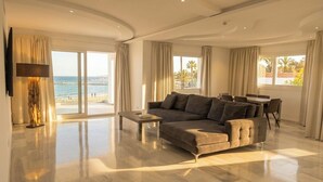 A spacious living room with a large sofa, a coffee table, and a TV mounted on the wall. There are large windows with curtains, a dining area with a table and chairs, and a view of the beach. - B51 Executive Flats Marbella Puerto Banus (Marbella)