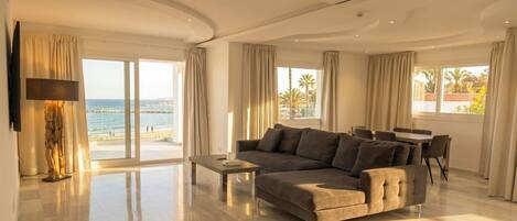 A spacious living room with a large sofa, a coffee table, and a TV mounted on the wall. There are large windows with curtains, a dining area with a table and chairs, and a view of the beach.