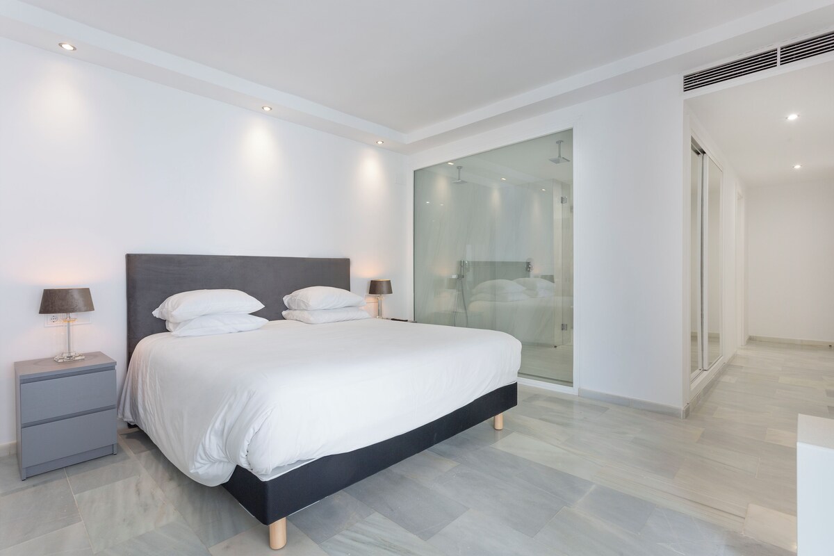 A modern bedroom with a large bed, bedside tables, and a glass shower enclosure.