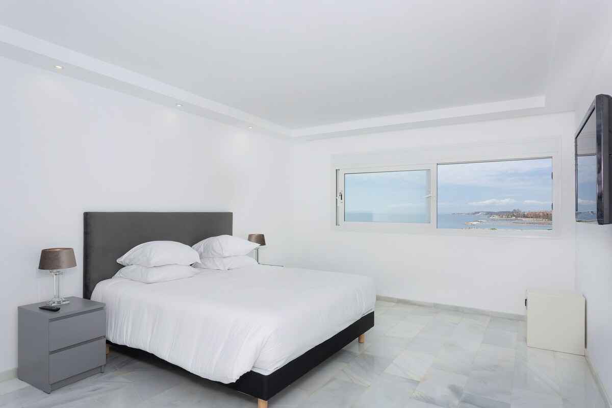 A modern bedroom with a large bed, a nightstand, a TV mounted on the wall, and a window with a coastal view.