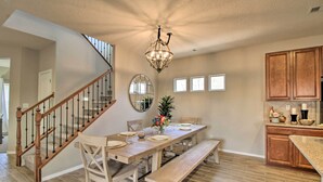Un comedor con mesa de madera, sillas y un banco. Se observa una escalera con barandillas de madera y un candelabro colgante.