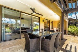 A modern outdoor dining area with a glass table and wicker chairs, a ceiling fan, and a view of the garden through glass doors.