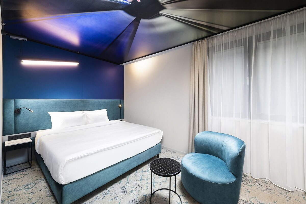 A hotel room with a bed, a blue chair, a small table, a ceiling fan, and a window with sheer curtains.