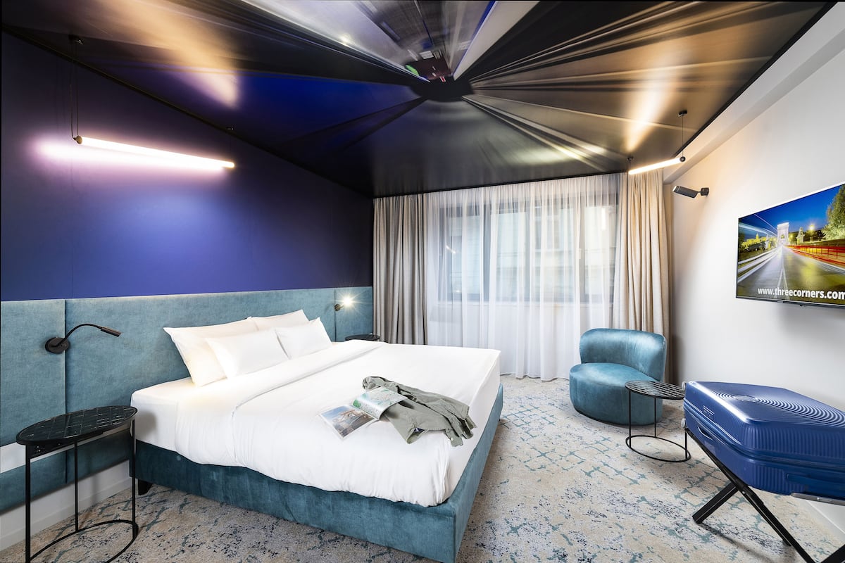 Une chambre d’hôtel moderne avec un grand lit, un mur d’accent bleu, un coin salon avec un fauteuil et une grande fenêtre avec des rideaux.