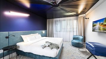 A modern hotel room with a large bed, a blue accent wall, a seating area with a chair, and a large window with curtains.
