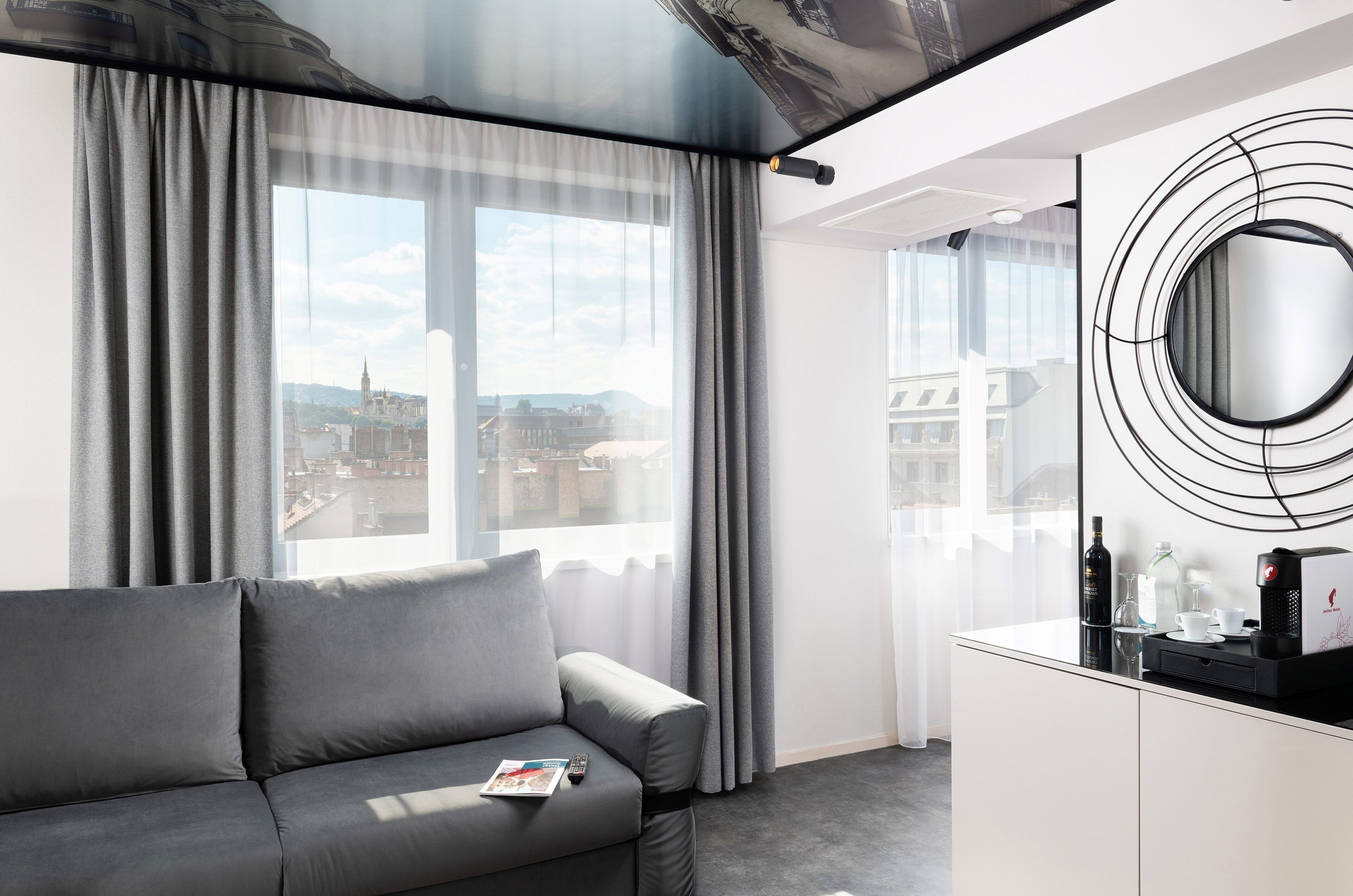 Habitación de hotel moderna con sofá gris, un ventanal con cortinas, un espejo redondo y una nevera con cafetera.