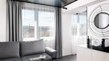 Habitación de hotel moderna con sofá gris, un ventanal con cortinas, un espejo redondo y una nevera con cafetera.