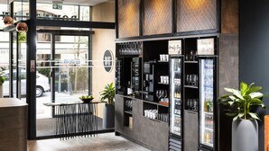 A modern bar area with a glass door entrance, a seating area with tables and chairs, and shelves stocked with glasses and bottles. - Three Corners Downtown Hotel (Budapest)