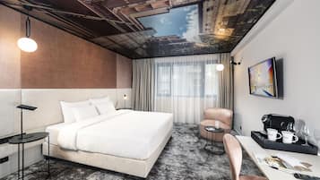 A modern hotel room with a large bed, a desk with a coffee maker, a chair, and a view of the sky through a ceiling mural.