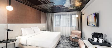 Una habitación de hotel moderna con una cama grande, un escritorio con cafetera, una silla y vistas al cielo a través de un mural en el techo.