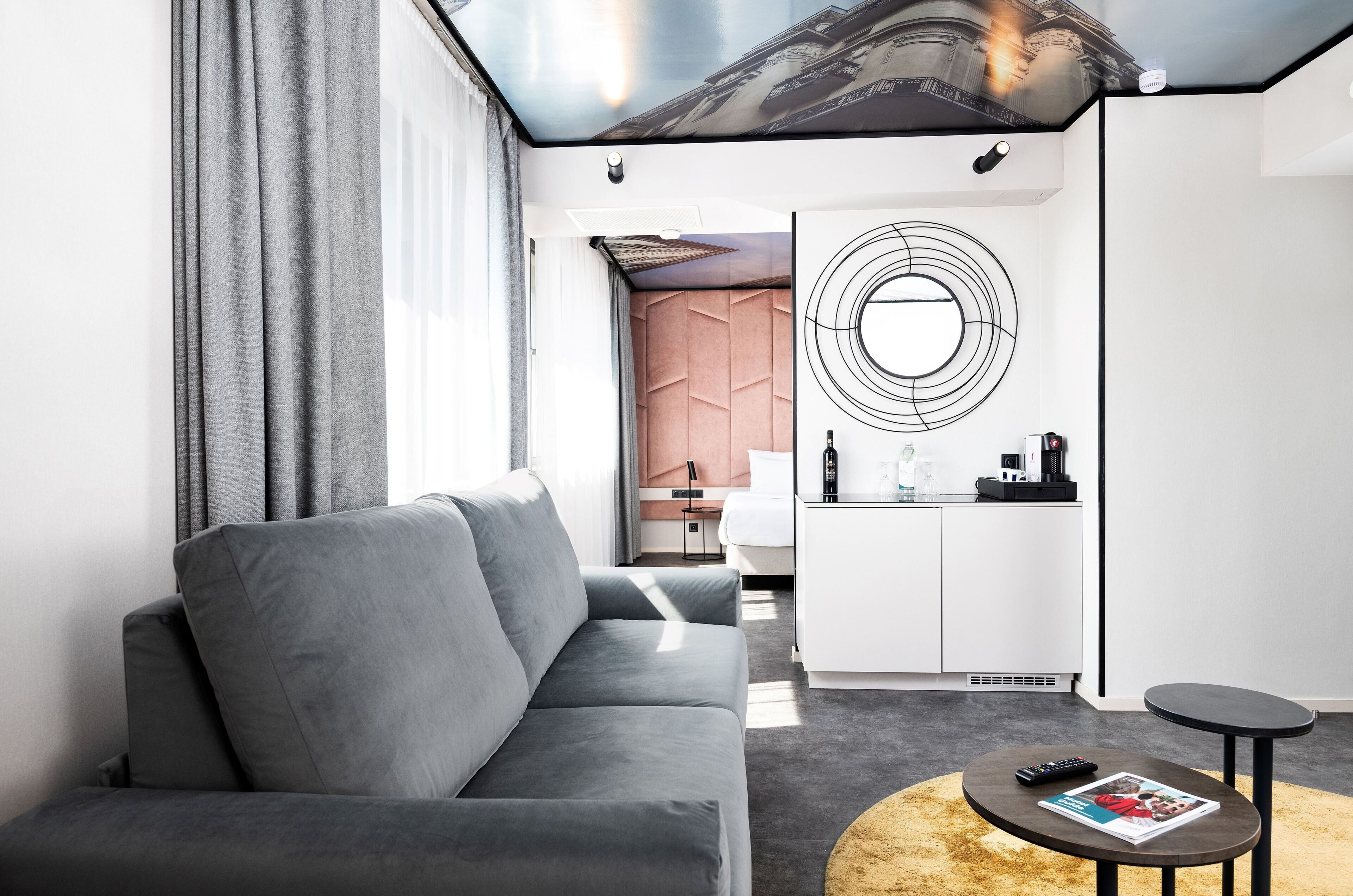 A modern hotel room with a grey sofa, a round coffee table, a kitchen area, and a decorative wall piece.