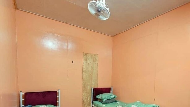 A small, single-room accommodation with two beds, a ceiling fan, and a patterned floor.